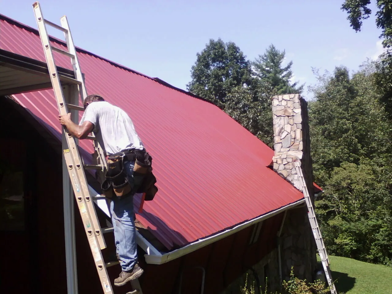 Roofer on a ladder beside a red metal roof and stone chimney — Soffit & Fascia Repair by Roofing Brothers in Culdesac